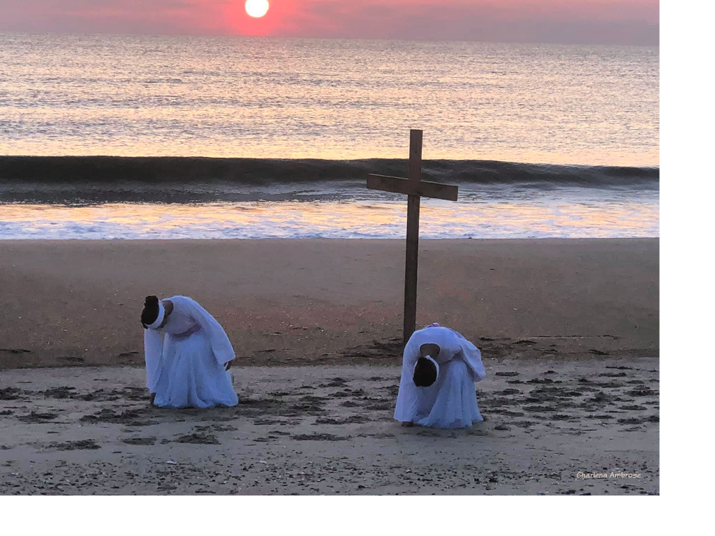 Early sunrise on Easter Morning at the ocean. Two girls dressed in a white long sleeve top and flowing skirt with light purple sash are dancing in front of a simple wooden cross. The dancers are bowed down facing the audience in an attitude of worship.