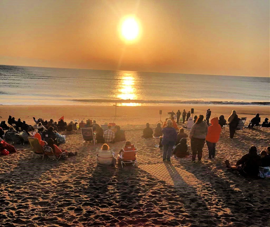 Early Easter Sunday morning on the beach. People have gathered to attend the Easter Son Rise Celebration. The sun has risen is shining brightly upon the water casting a beautiful reflection.