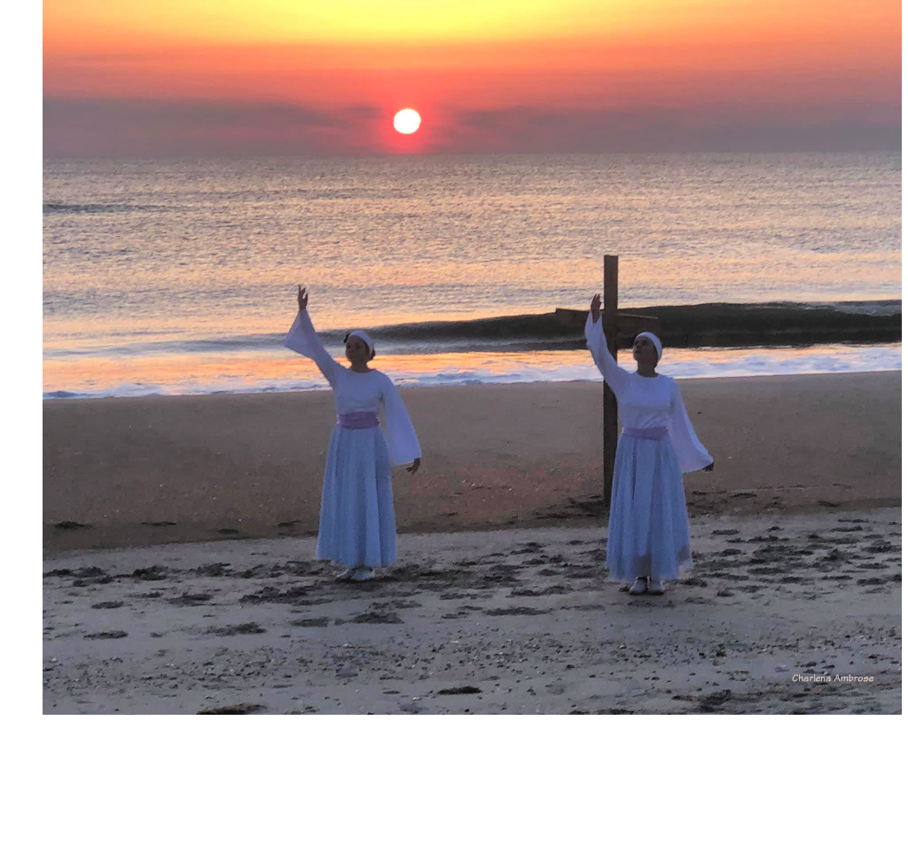 Early sunrise on Easter Morning at the ocean. Two girls dressed in a white long sleeve top and flowing skirt with light purple sash are dancing in front of a simple wooden cross. The dancers have one arm lifted upwards pointing towards towards heaven while the other arm is down by their side.