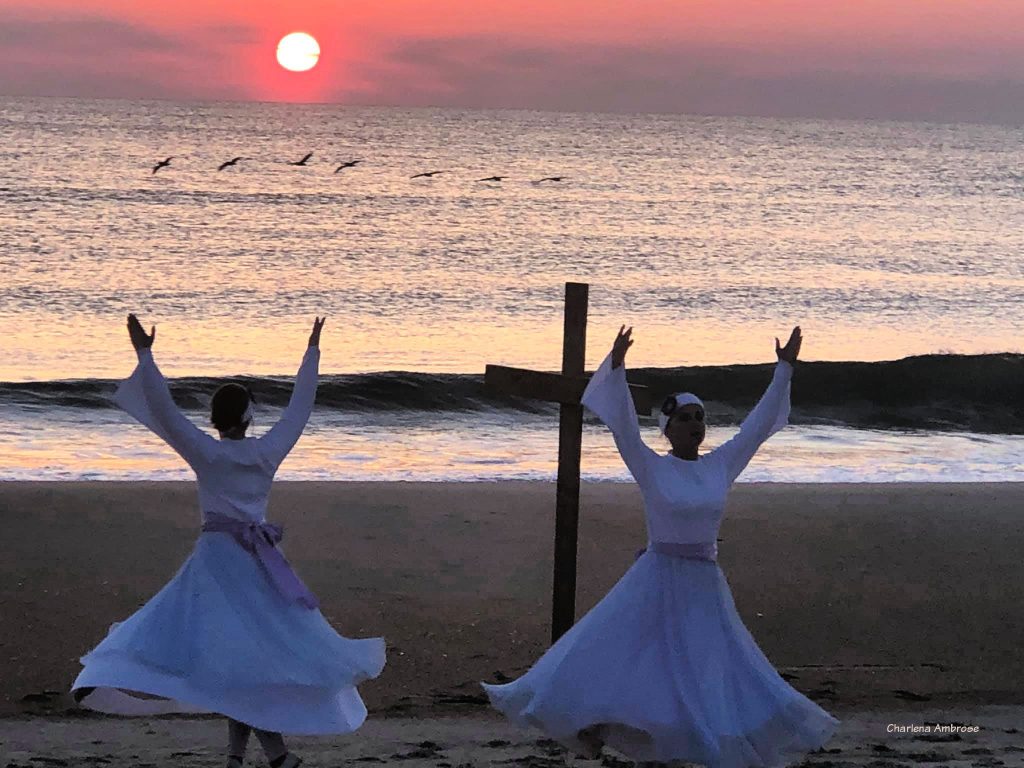 Early sunrise on Easter morning at the ocean. Two girls dressed in a white long sleeve top and flowing skirt with light purple sash are dancing in front of a simple wooden cross. The dancers have their arms lifted upwards in an attitude of praise as their billowy skirts twirl about their legs.