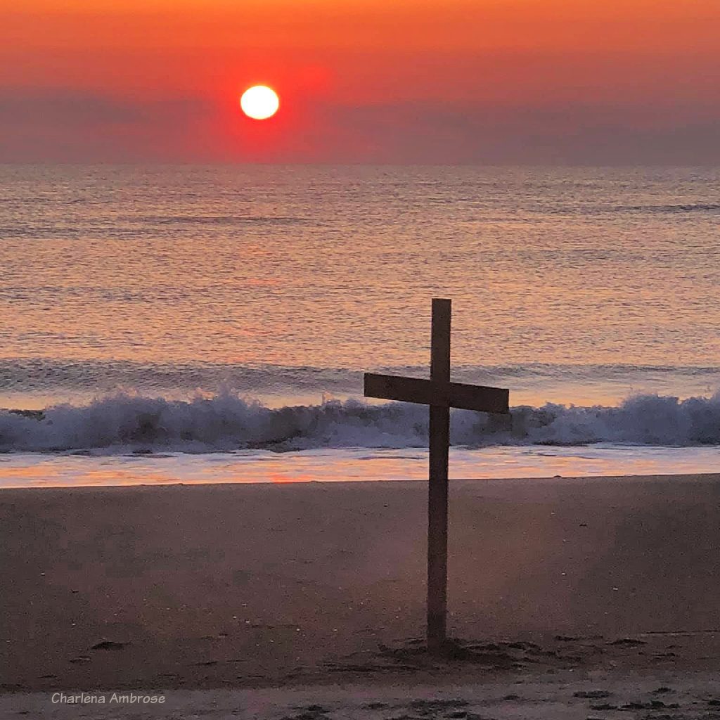 Easter Sunday Morning sunrise on the beach. the sky is filled with warm beautiful oranges of the sun as it is rising above the ocean. A simple Wooden cross has been erected at the shoreline on a calm morning.