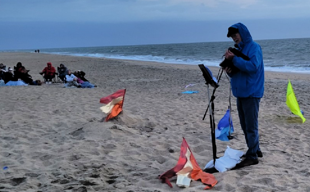 Early cold morning at the beach. The minister is dressed in a blue winter coat and is standing at the microphone preaching. There are 2 small flags on the ground in front of him. They are gold, orange and red. Audience members are seated in the background.
