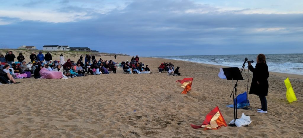 Early morning at the ocean. A crown has gathered for Easter sunrise celebration. A female singer is at the microphone facing the audience. Everyone is dressed in coats for the early cold morning.